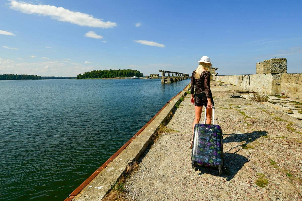 Seorang wanita berjalan di tepi laut dengan menarik koper bermotif warna-warni di bawah langit cerah dan pemandangan pulau hijau di kejauhan.