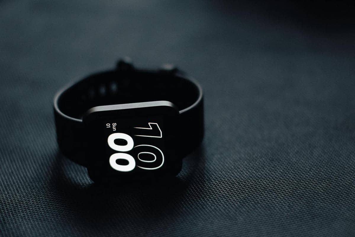 Smartwatch hitam dengan layar digital menampilkan waktu 10:00 terletak di permukaan gelap bertekstur.