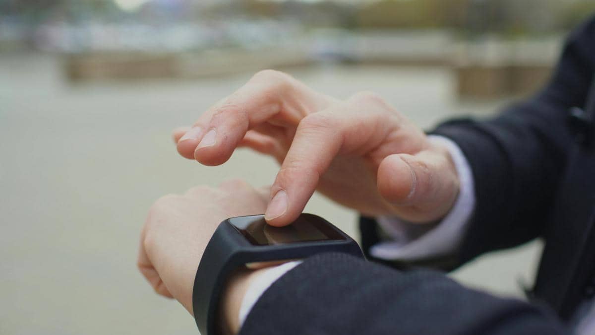 Seseorang mengenakan jas menyentuh layar smartwatch hitam di pergelangan tangan saat berada di luar ruangan.