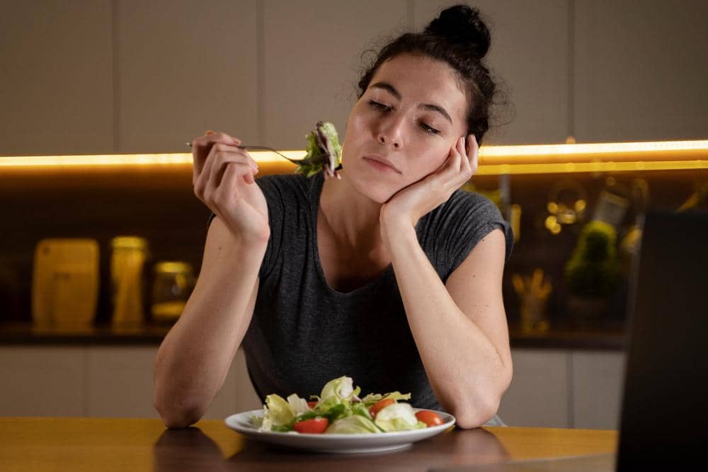 Seorang perempuan duduk di meja makan menatap piring salad dengan ekspresi lesu, tampak kehilangan nafsu makan di dapur modern.