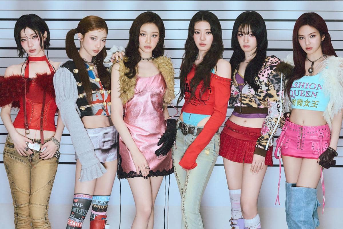 Enam anggota grup musik perempuan berpose bersama mengenakan busana warna-warni bergaya Y2K di depan latar bergaris hitam putih.