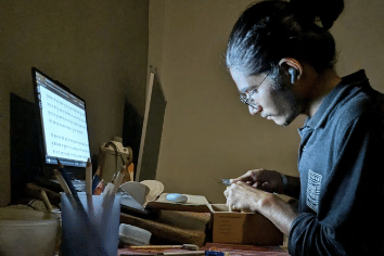 Seorang pria bekerja di meja dengan komputer dan perlengkapan kerja di malam hari, fokus pada pekerjaan sampingan di rumah.