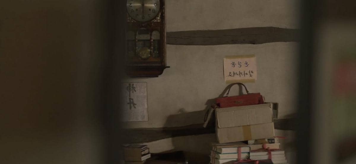 Tumpukan buku dan kotak kardus di dalam ruangan bergaya tradisional dengan jam dinding tua dan tulisan aksara Korea di dinding.