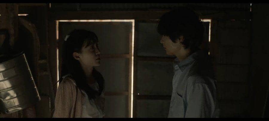 Dua karakter berdiri saling berhadapan di ruangan gelap dengan cahaya redup dari jendela kayu dalam adegan drama The Scarecrow.