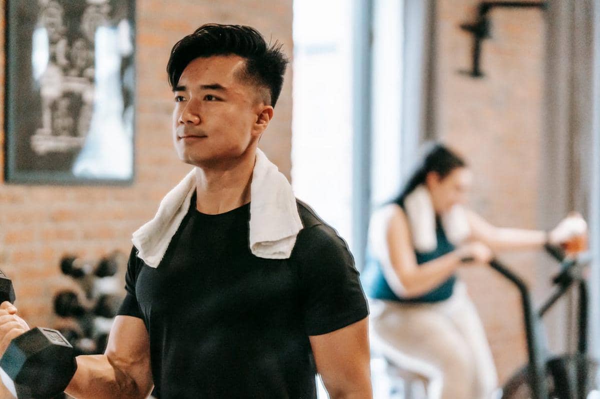 Pria mengenakan kaus hitam berolahraga dengan dumbbell di gym, handuk putih di leher, dengan latar belakang wanita bersepeda statis.