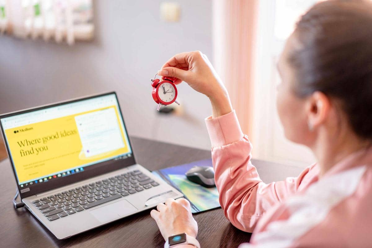 Seseorang mengenakan pakaian merah muda memegang jam kecil berwarna merah sambil duduk di depan laptop yang terbuka di meja kerja.