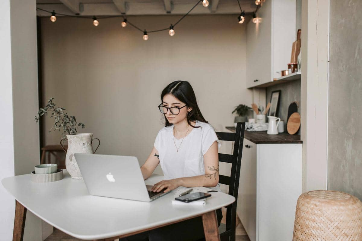 Seorang wanita berkacamata bekerja dengan laptop di meja putih dalam ruangan bergaya minimalis dengan lampu gantung dekoratif.
