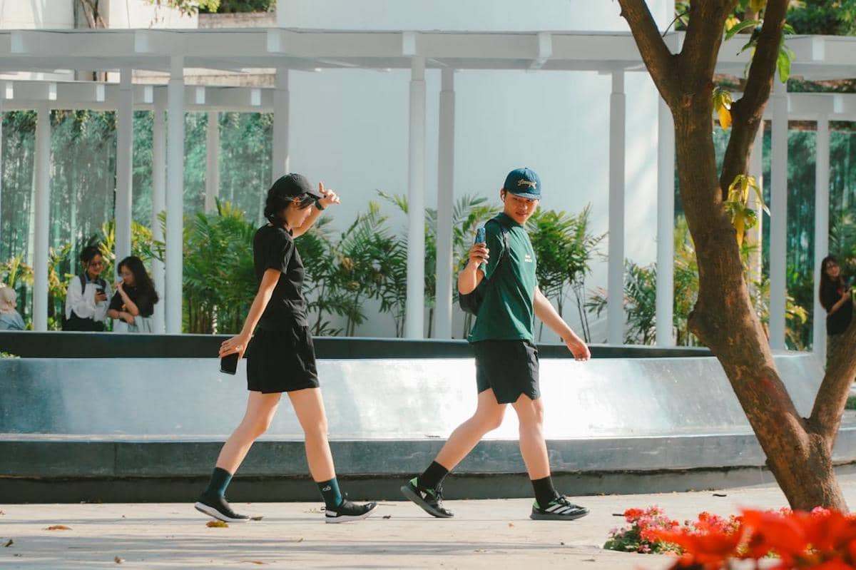Dua orang berjalan santai di taman kota dengan latar pepohonan dan bangunan putih di siang hari yang cerah.