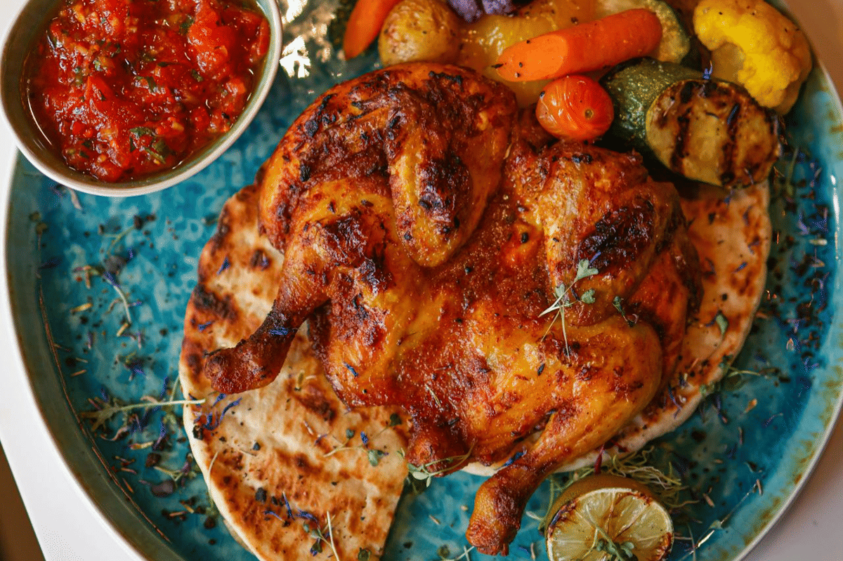 Hidangan pollo alla diavola berupa ayam panggang berbumbu pedas disajikan di atas roti pipih dengan sayuran panggang dan saus tomat.