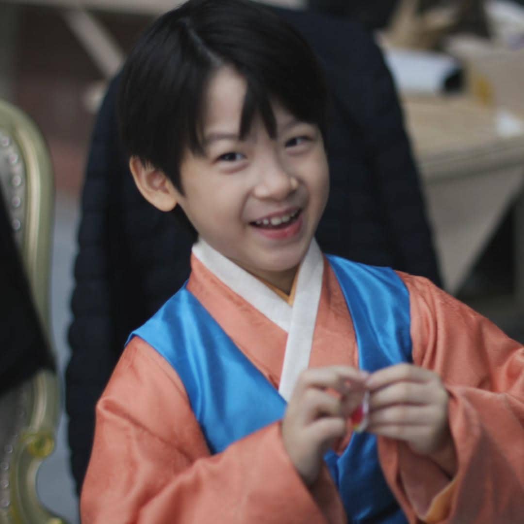 Anak laki-laki tersenyum mengenakan hanbok berwarna oranye dan biru di lokasi syuting drama Perfect Crown.