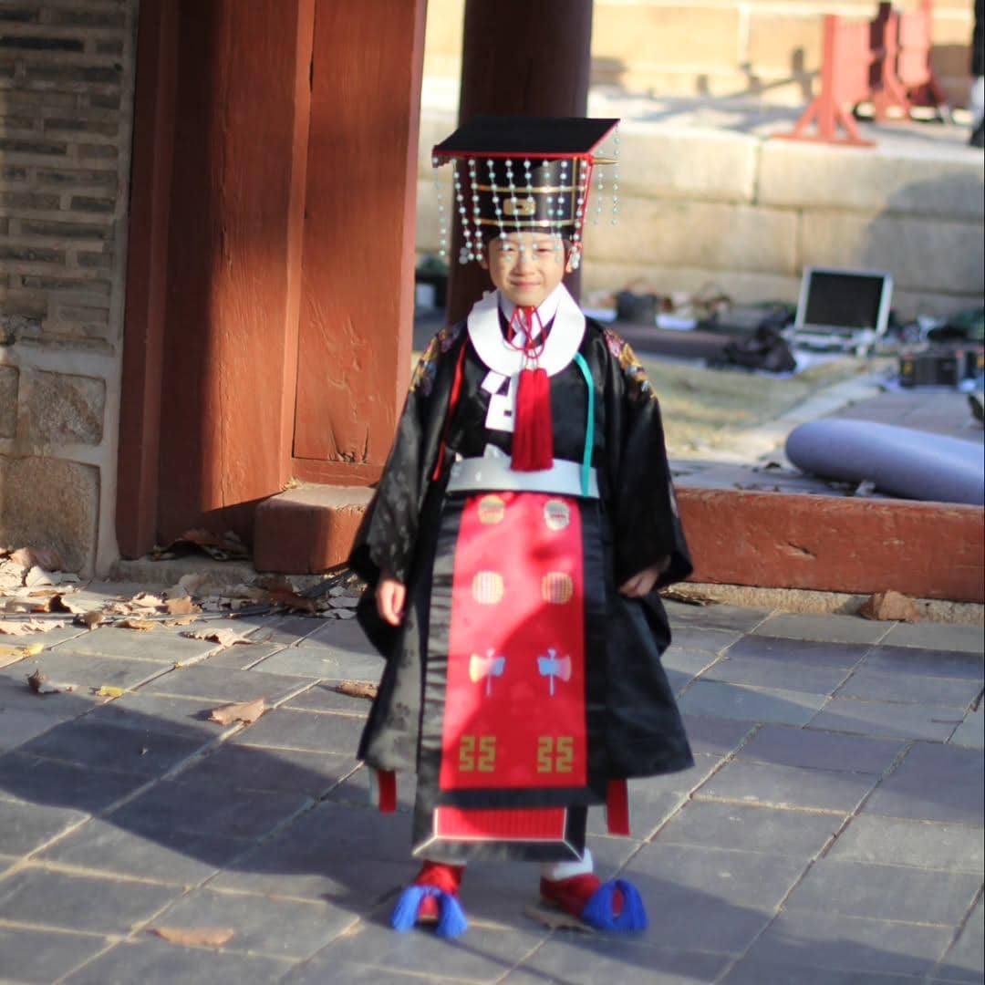 Seorang anak mengenakan hanbok tradisional Korea lengkap dengan hiasan kepala kerajaan di area berarsitektur kayu dan batu.