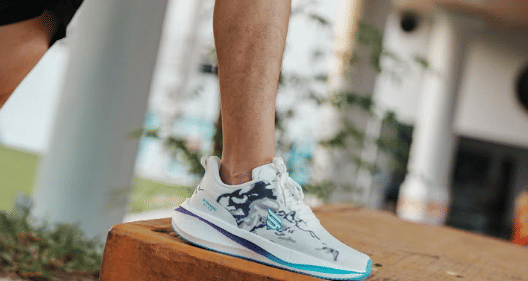 Seseorang mengenakan sepatu olahraga Stradenine berwarna putih dengan aksen biru dan ungu saat berada di luar ruangan.