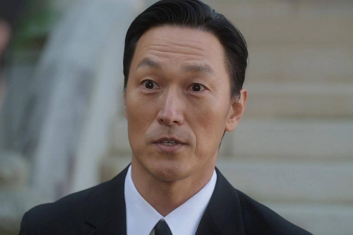 Seorang pria mengenakan setelan jas hitam dan dasi hitam berdiri dengan ekspresi serius dalam adegan drama Korea Perfect Crown.