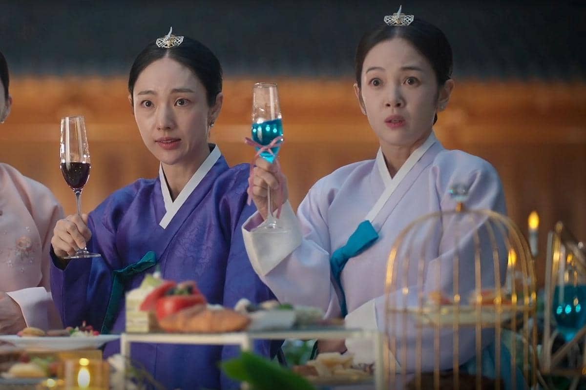 Dua wanita mengenakan hanbok tradisional Korea berwarna ungu dan biru muda memegang gelas minuman di meja penuh hidangan.