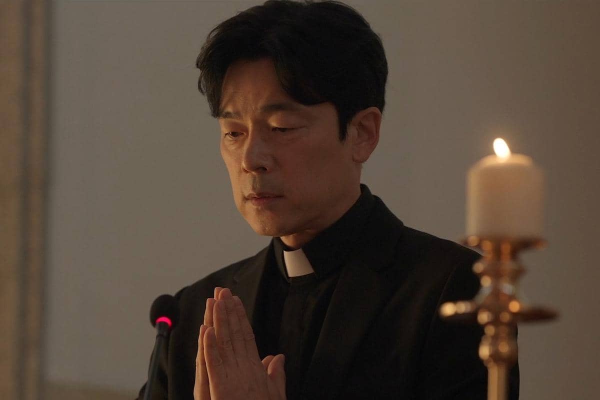 Seorang pria berpakaian hitam dengan kerah pendeta sedang berdoa di depan lilin menyala dalam adegan drama Korea Perfect Crown.