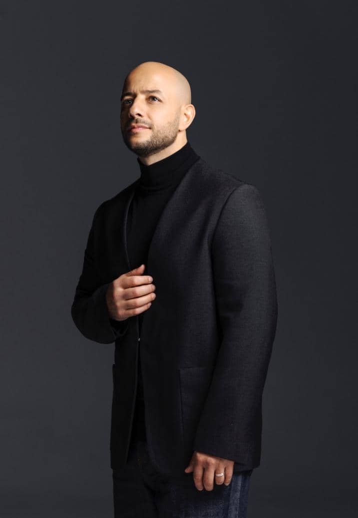 Seorang pria berkepala plontos mengenakan jas gelap dan turtleneck hitam berdiri dengan latar belakang abu-abu gelap.