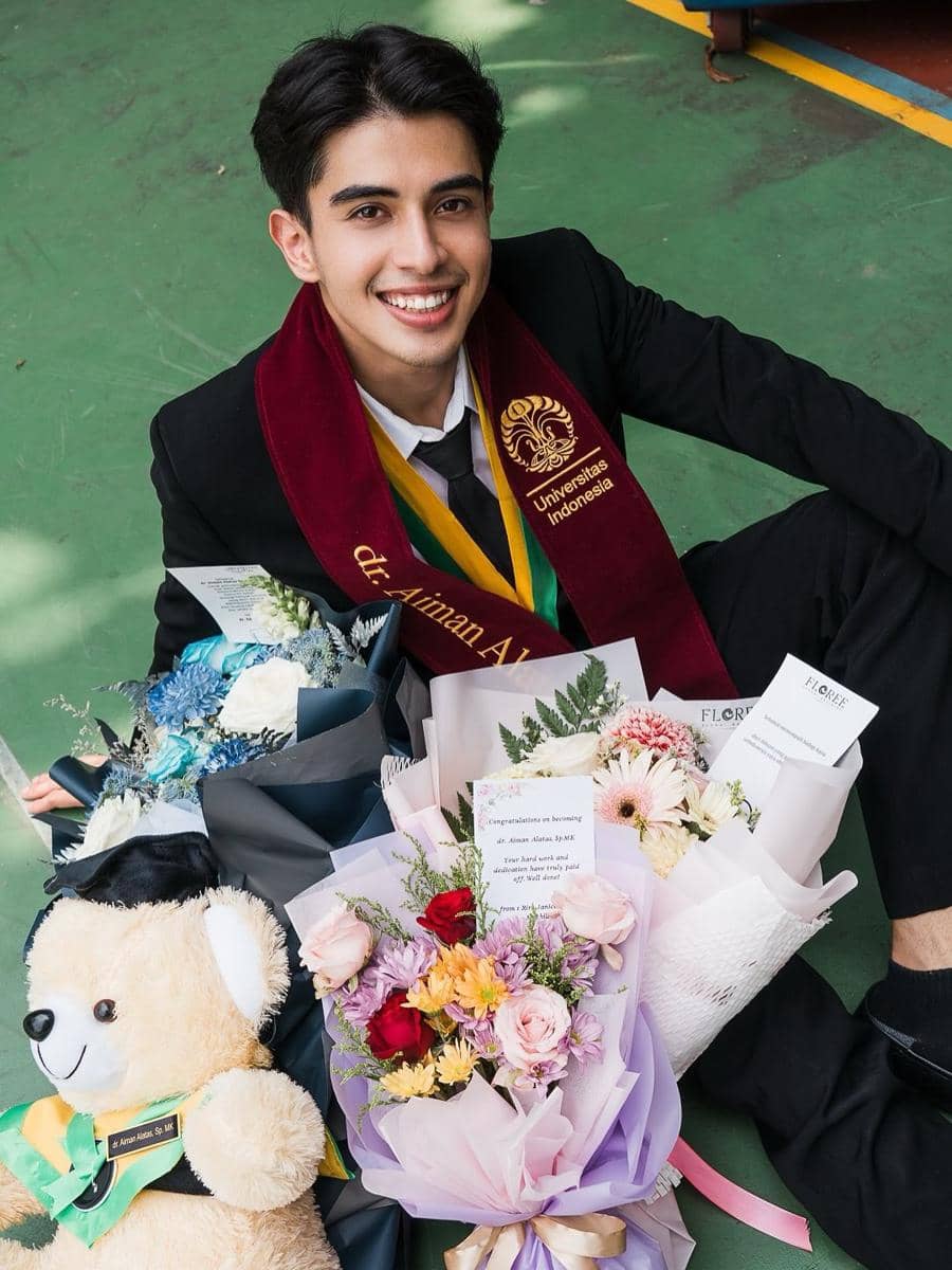 Seorang pria mengenakan jas hitam dan selempang Universitas Indonesia tersenyum sambil duduk di lantai dengan buket bunga dan boneka hadiah wisuda di sekitarnya.