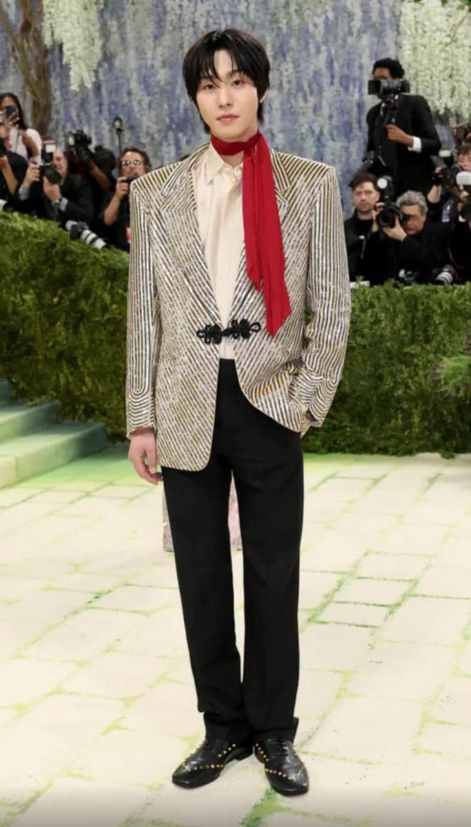 Seorang pria mengenakan jas bergaris emas, kemeja krem, celana hitam, dan syal merah di acara Met Gala dengan latar fotografer.