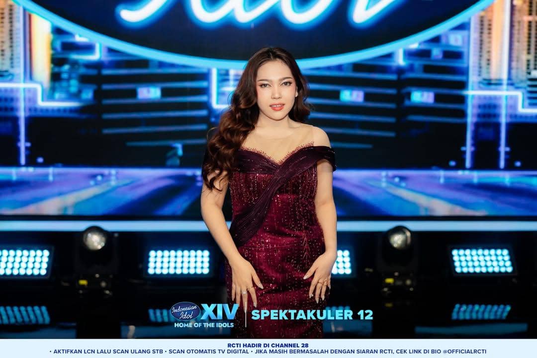 Seorang kontestan Indonesian Idol Season 14 mengenakan gaun merah marun berkilau berdiri di panggung dengan latar logo Indonesian Idol.