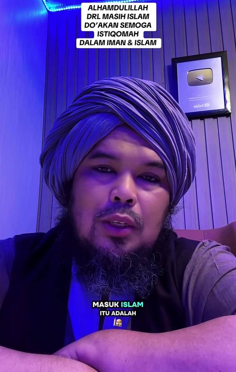 Seorang pria berjanggut mengenakan sorban abu-abu duduk di ruangan bernuansa biru dengan tulisan motivasi tentang keimanan di latar belakang.