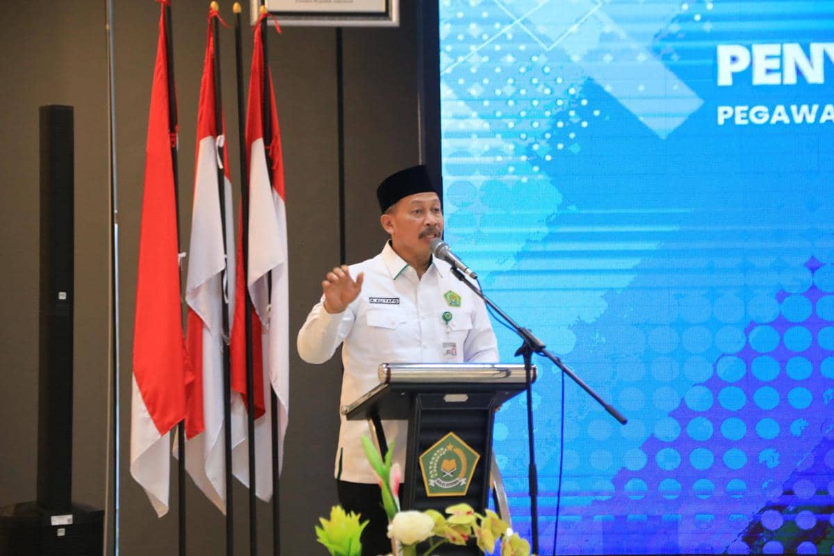 Seorang pejabat Kementerian Agama Sulawesi Selatan berbicara di podium dengan latar layar biru dan bendera Indonesia di sampingnya.