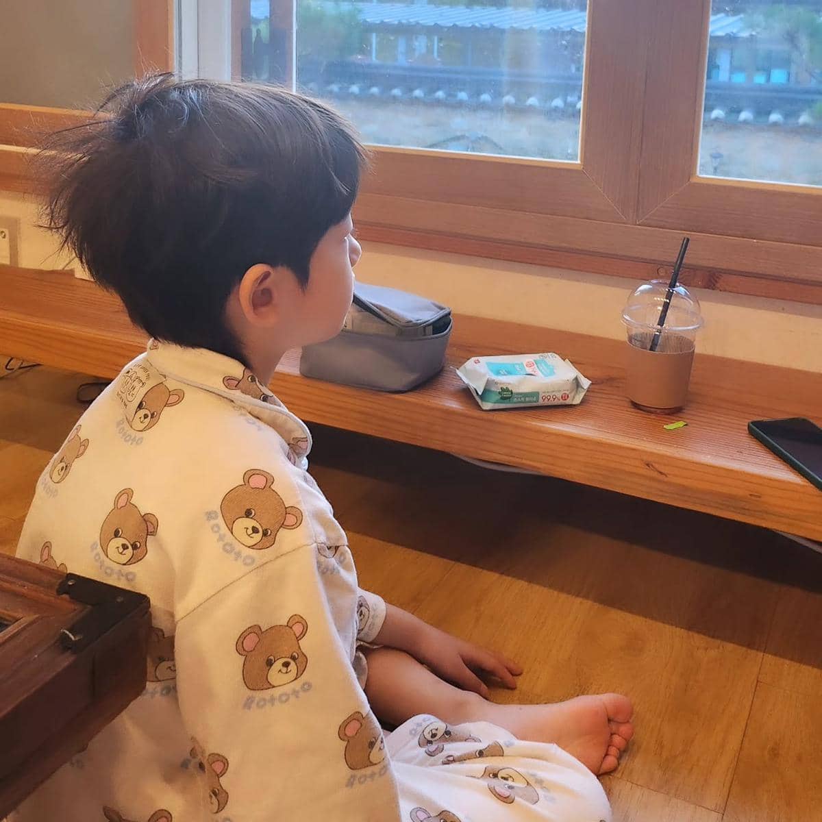 Seorang anak kecil mengenakan piyama bergambar beruang duduk di lantai kayu sambil memandang keluar jendela di ruangan tradisional.