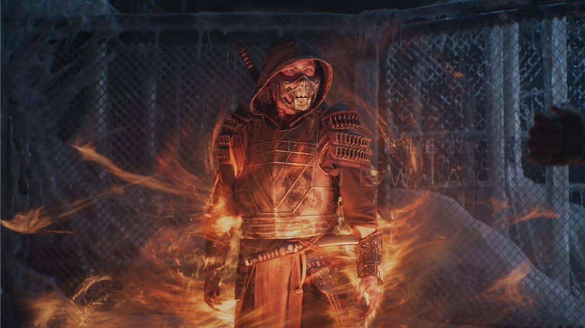 Karakter bertopeng dengan baju zirah dan tudung berdiri dikelilingi api dalam adegan film Mortal Kombat.