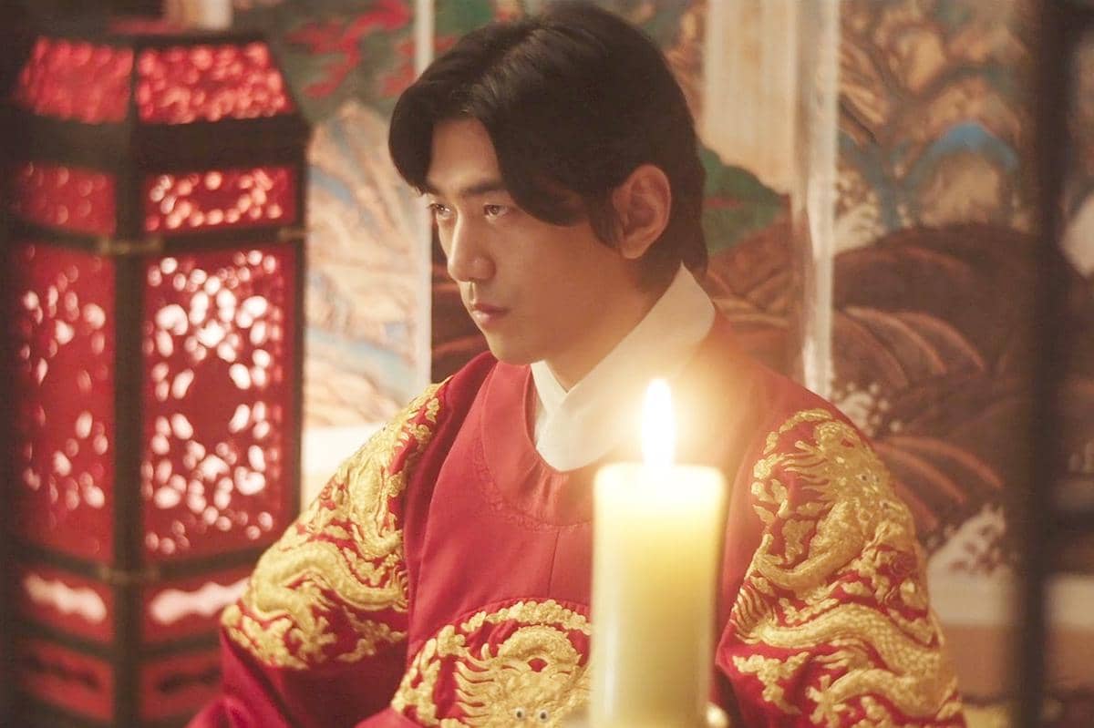 Seorang pria mengenakan hanbok kerajaan berwarna merah dengan bordiran naga emas duduk di depan lilin menyala dalam adegan drama Korea Perfect Crown.