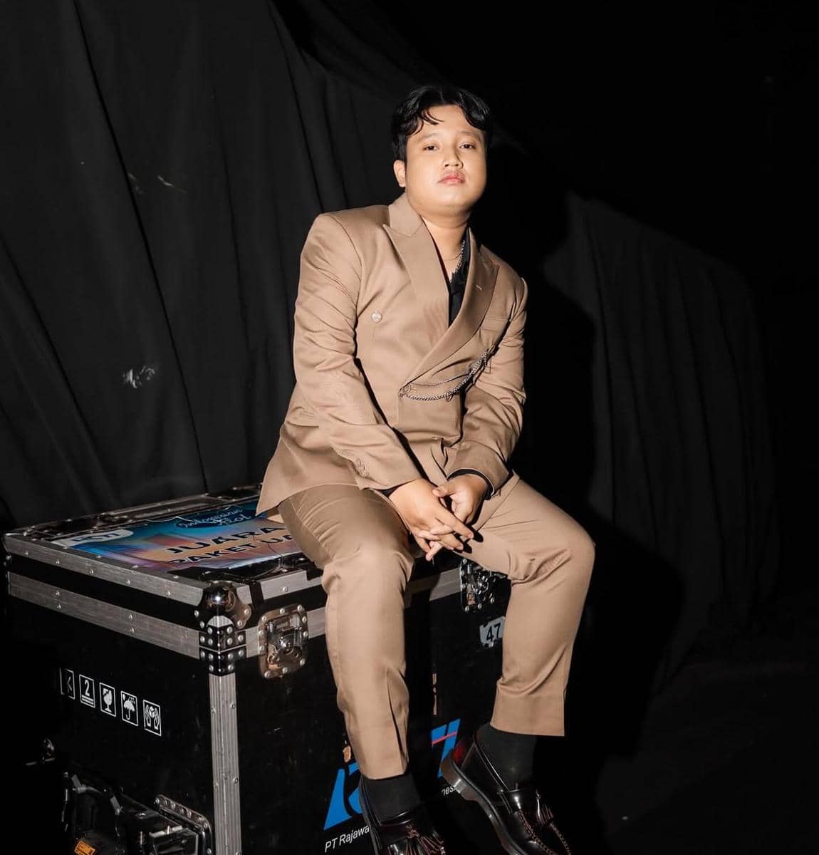 Seorang pria mengenakan setelan jas cokelat duduk di atas peti peralatan dengan latar belakang hitam dalam pencahayaan studio.