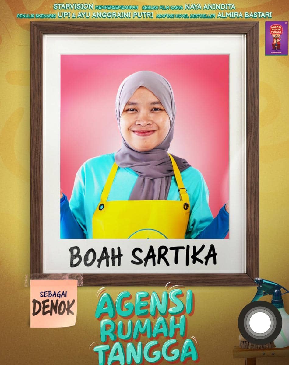 Boah Sartika mengenakan hijab abu-abu dan celemek kuning berpose di depan latar merah muda sebagai karakter Denok dalam film Agensi Rumah Tangga.