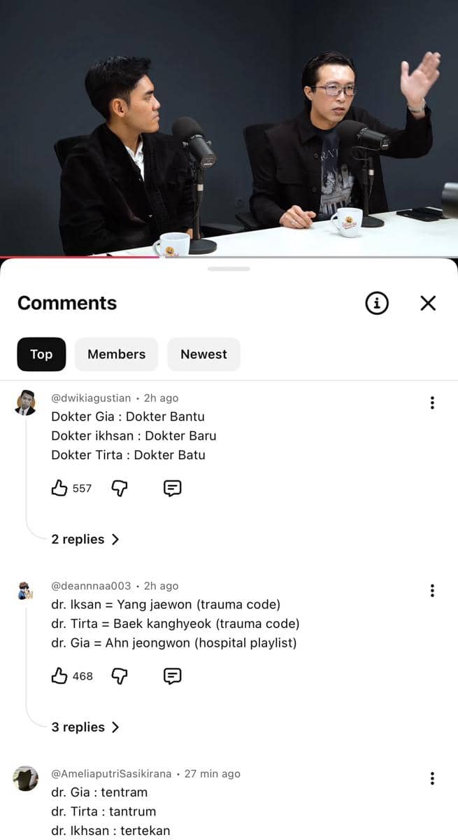 Dua pria duduk di studio podcast dengan mikrofon di depan mereka, berbicara serius sambil memegang cangkir kopi di meja putih.