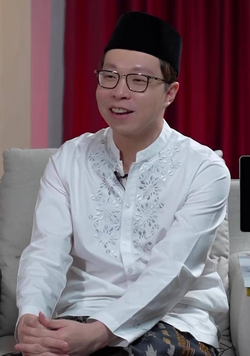 Seorang pria berkacamata mengenakan baju koko putih bersulam dan peci hitam sedang duduk tersenyum di sofa dengan latar tirai merah.