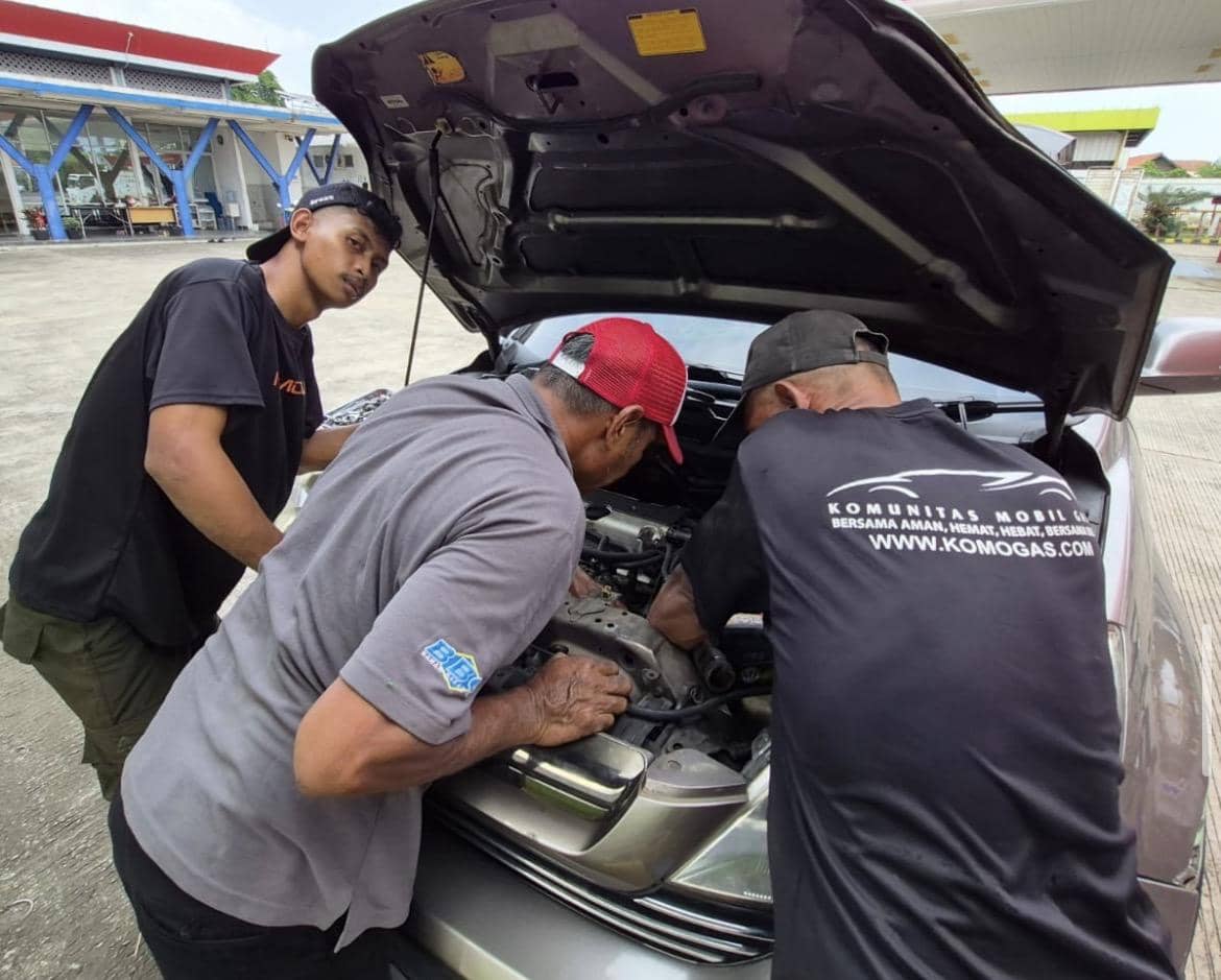 Tiga orang mekanik sedang memeriksa mesin mobil dengan kap terbuka di area luar bengkel untuk konversi bahan bakar gas.