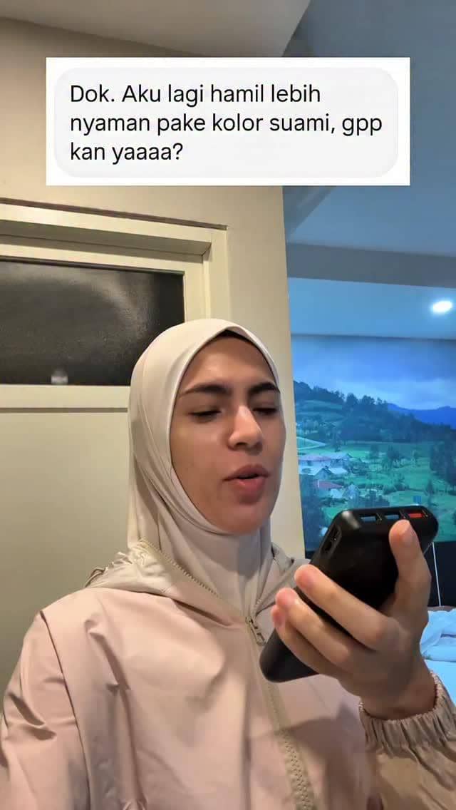 Seorang wanita berhijab mengenakan jaket krem sedang memegang ponsel dan membaca pesan tentang kenyamanan pakaian saat hamil.