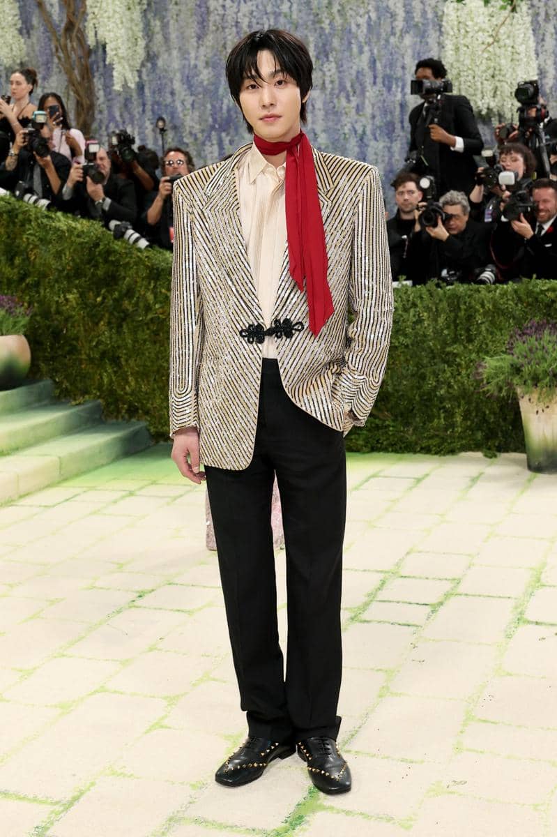 Seorang pria mengenakan jas bergaris emas, kemeja krem, dan syal merah berpose di karpet acara Met Gala dengan latar fotografer.