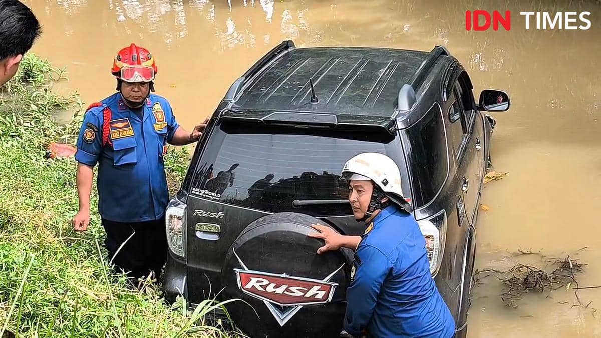 Petugas berseragam biru berhelm mengevakuasi minibus hitam yang terperosok ke dalam parit berlumpur di area Bandara Soetta.
