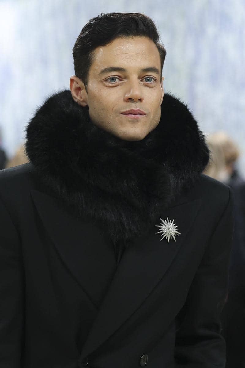 Rami Malek mengenakan jas hitam dengan kerah bulu besar dan bros berbentuk bintang di dada saat menghadiri acara Met Gala 2026.