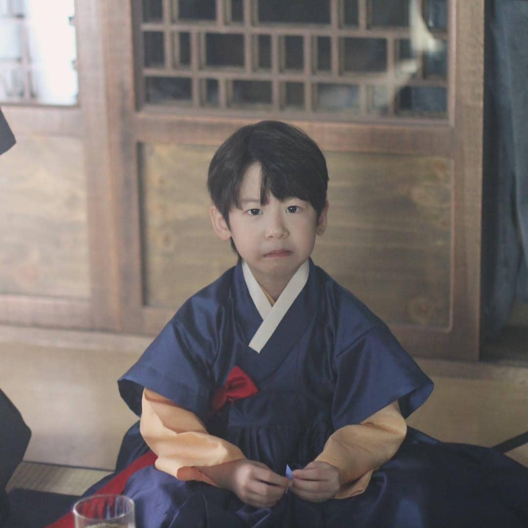 Seorang anak laki-laki mengenakan hanbok biru dan duduk di dalam ruangan tradisional Korea dengan ekspresi serius di wajahnya.
