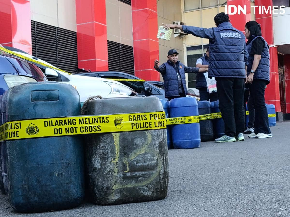 Petugas kepolisian mengenakan rompi reskrim berdiri di dekat deretan jeriken BBM yang disita, dibatasi garis polisi di depan gedung berwarna merah.