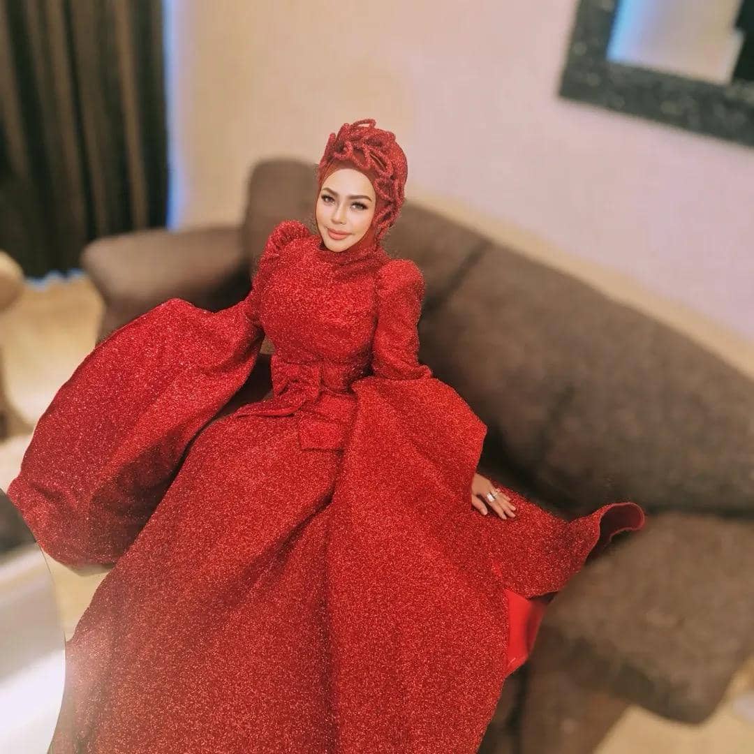 Seorang wanita mengenakan gaun merah berkilau dengan hijab senada, duduk di sofa berwarna cokelat di dalam ruangan.