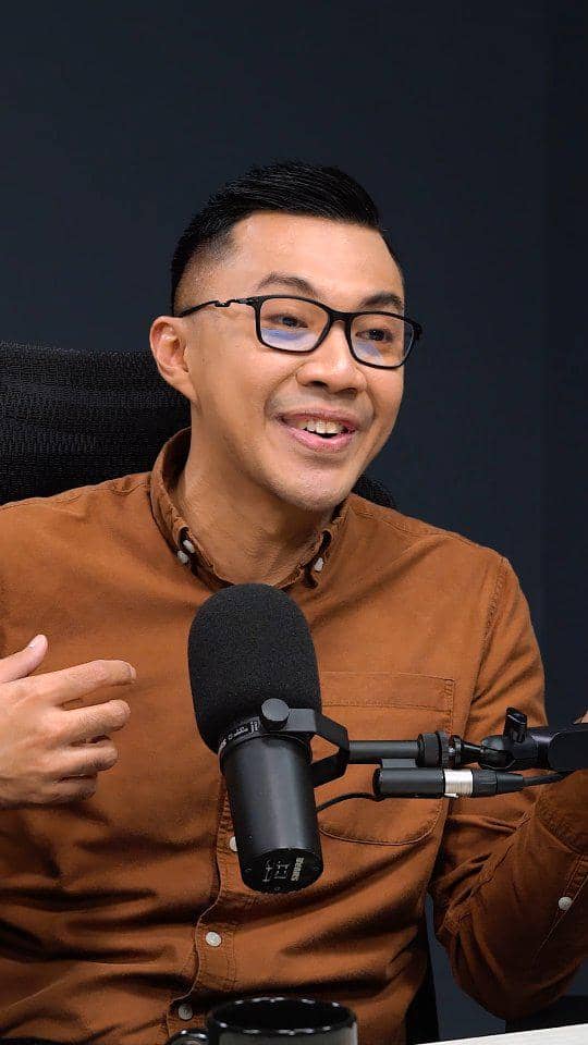 Seorang pria berkacamata mengenakan kemeja cokelat sedang berbicara di depan mikrofon dalam suasana rekaman podcast.