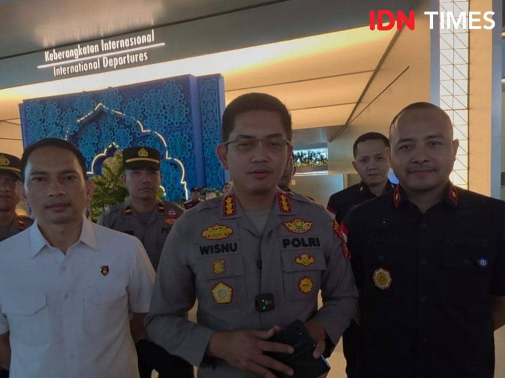 Beberapa petugas kepolisian dan imigrasi berdiri di area keberangkatan internasional Bandara Soekarno-Hatta dengan latar dekorasi biru.