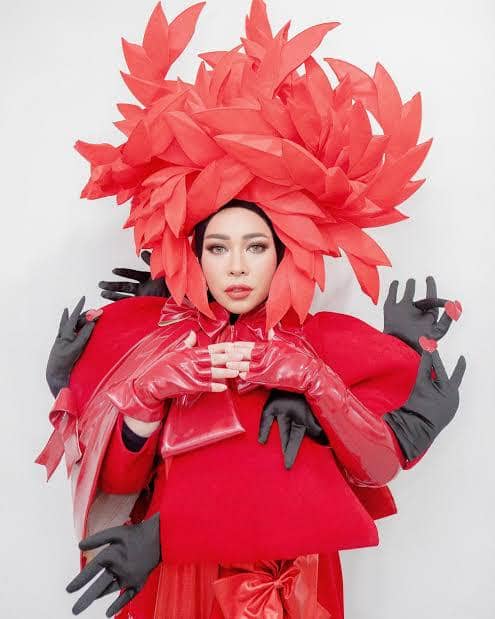 Seorang perempuan mengenakan kostum merah mencolok dengan hiasan kepala besar berbentuk daun dan beberapa tangan berwarna hitam di sekelilingnya.