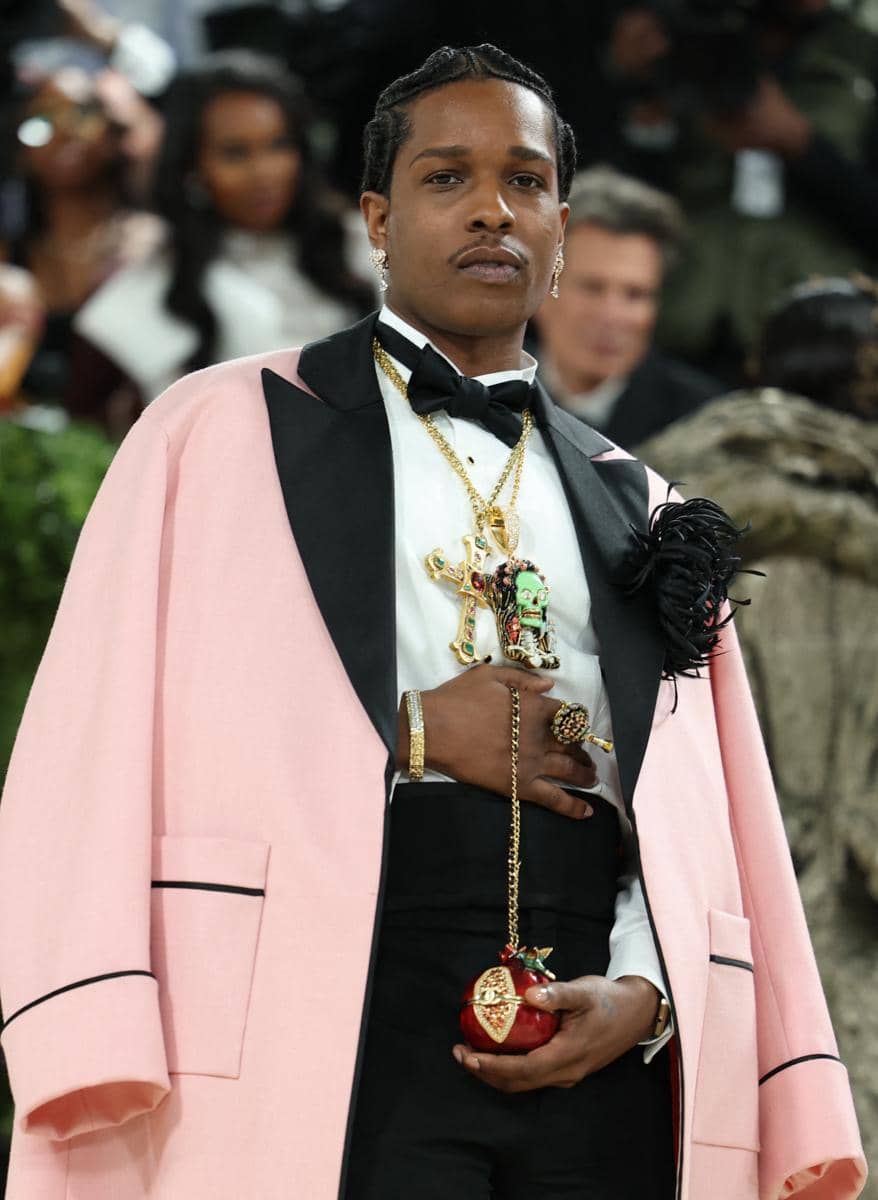 Seorang pria mengenakan jas merah muda dengan detail hitam, dasi kupu-kupu, dan perhiasan emas saat menghadiri acara Met Gala 2026.