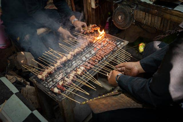 Beberapa orang memanggang sate kambing di atas bara arang dengan kipas angin kecil di dekatnya untuk menjaga api tetap menyala.