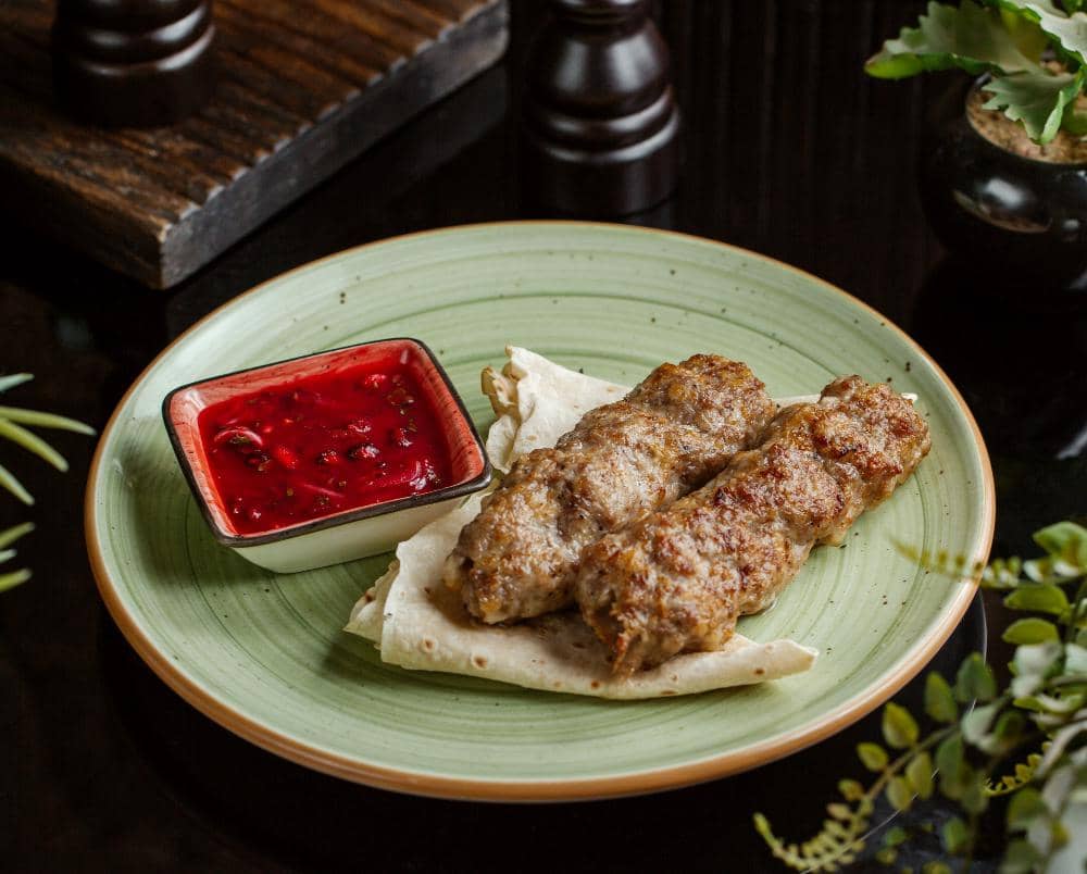 Dua potong cevapi panggang disajikan di atas roti pipih dengan saus merah dalam mangkuk kecil di piring hijau.