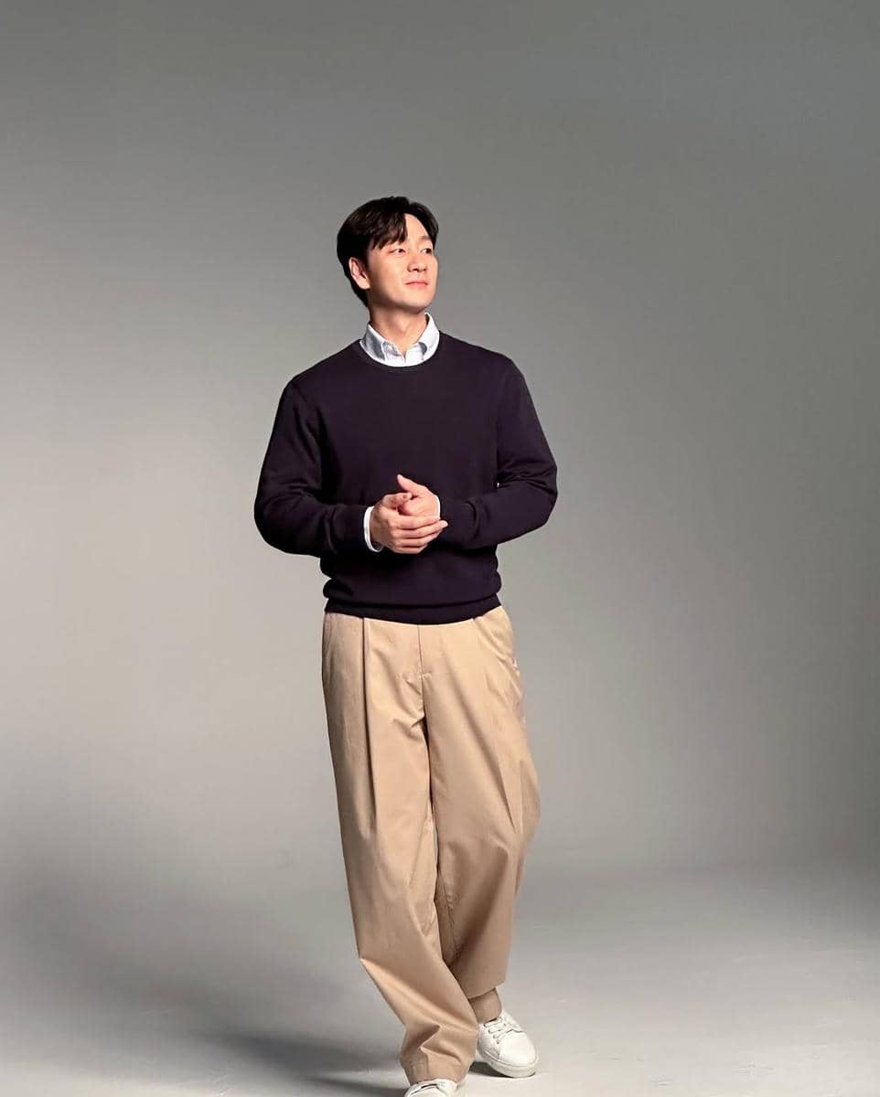Seorang pria bergaya kasual mengenakan sweater hitam, kemeja putih, celana krem longgar, dan sepatu putih di studio dengan latar abu-abu.