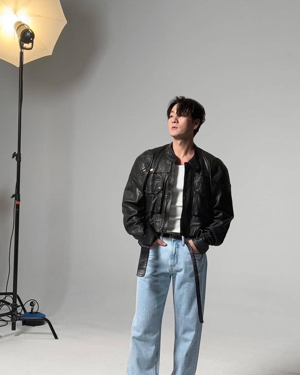 Seorang pria bergaya kasual mengenakan jaket kulit hitam, kaus putih, dan celana jeans longgar di studio dengan pencahayaan profesional.