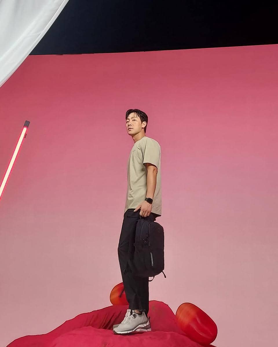 Seorang pria bergaya kasual mengenakan kaus krem, celana gelap, dan sepatu sneakers sambil memegang ransel di latar merah muda.