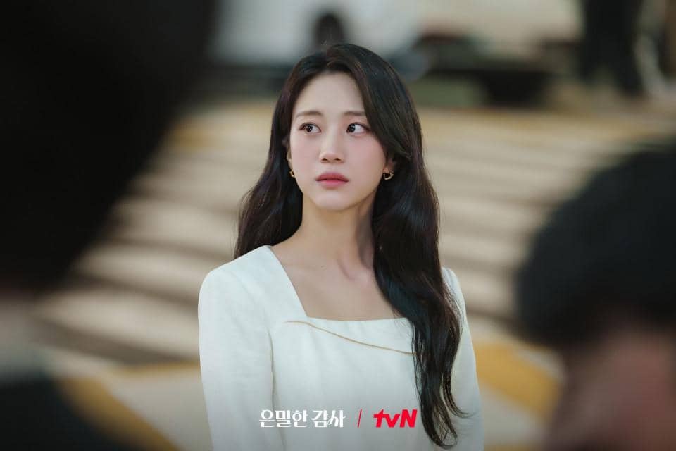 Seorang wanita berambut panjang mengenakan gaun putih berdiri di tempat umum dengan ekspresi serius dalam cuplikan drama Korea Filing for Love.